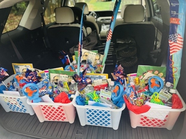 Four baskets filled with assorted snacks, toys, and patriotic decorations sit in the back of a car, ready to celebrate the military community. Each basket features red, white, and blue décor with pinwheels and American flag-themed items.