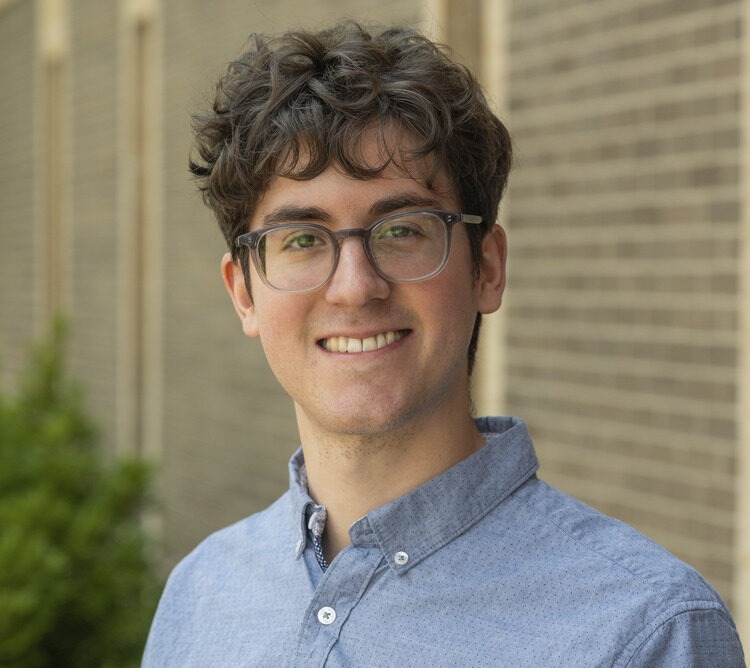 A young man with curly brown hair, glasses, and a blue button-up shirt smiles in front of a brick wall with greenery visible on the left.