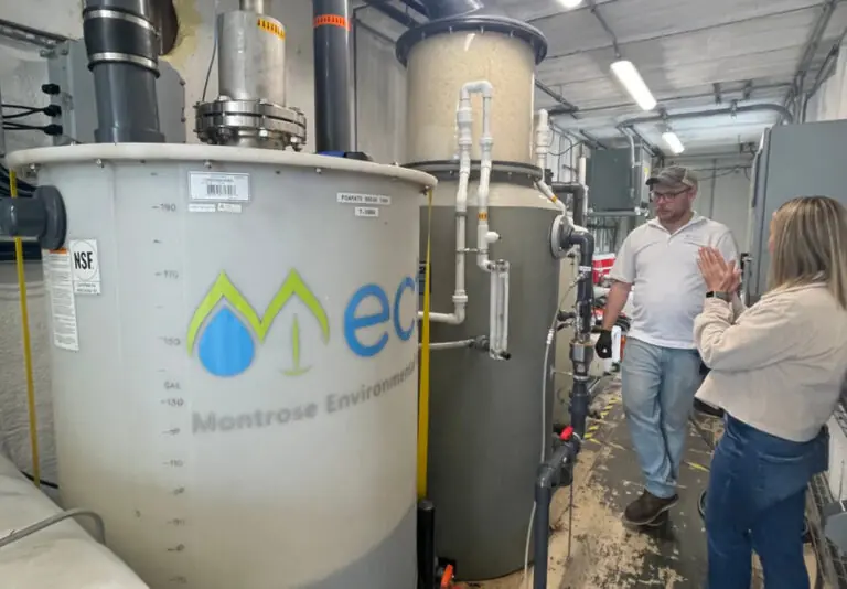 Two people stand in a facility next to large water treatment tanks labeled Montrose Environmental, discussing the equipment. The space is industrial, with pipes and control panels visible around them.