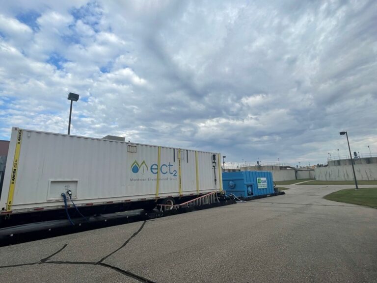 A white shipping container labeled “ect2” and a blue unit are set up on paved ground outdoors near a fence, under a cloudy sky.