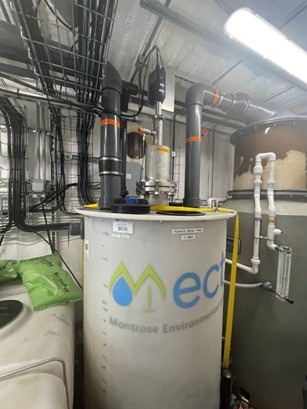 A large chemical tank labeled Montrose Environmental with attached pipes and wiring in an industrial setting under bright overhead lights, featured as part of a PFAS Pilot Story.