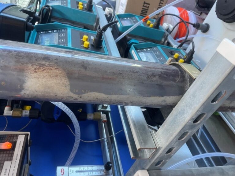 A close-up of a metal pipe mounted on a rack, with control panels, wires, and tubes in the background, suggesting an industrial or laboratory setup—an environment reminiscent of a PFAS Pilot Story.