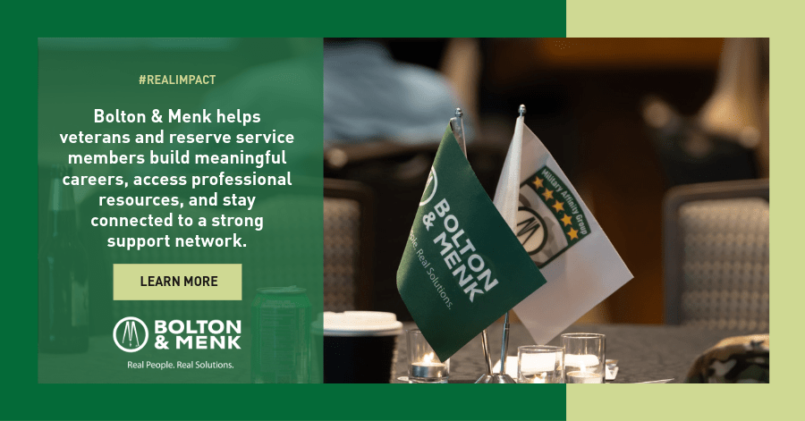 Two small table flags, one with the Bolton & Menk logo and one with a military emblem, sit next to brochures on a table. Text highlights Bolton & Menk’s support for veterans and service members.