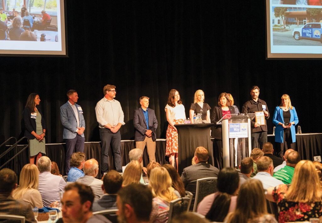 Nine people stand on stage at a United Way event, some at a podium, speaking to an audience. Projected images are visible above them. The audience is seated and facing the stage.