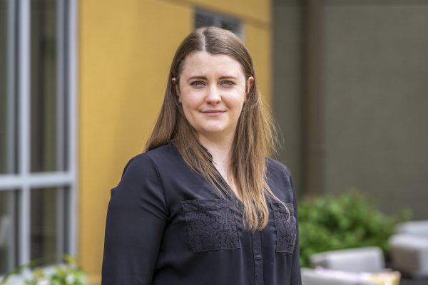 A woman with long brown hair wearing a black blouse stands outdoors in front of a yellow and gray building, looking at the camera with a neutral expression. There is greenery and outdoor furniture in the background.