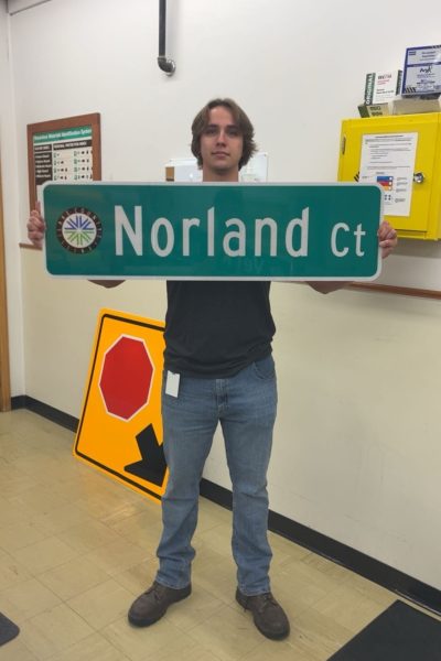 A person in a gray shirt and jeans stands indoors holding a green street sign that reads Norland ct. Behind them, there is a yellow stop sign and a yellow wall-mounted box.