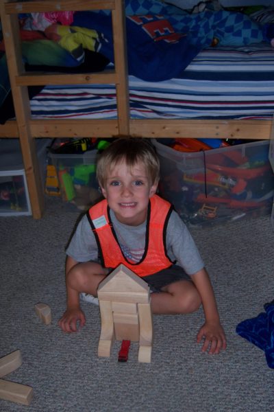 A young boy wearing an orange safety vest sits on the floor, smiling, with wooden building blocks arranged in front of him. Behind him is a bunk bed with colorful bedding and plastic bins of toys.