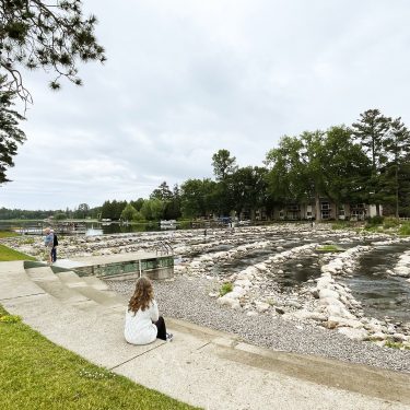 Image-10 A person with long hair sits on a curved concrete path by a river with rock formations, trees, and a few people walking in the background under an overcast sky.