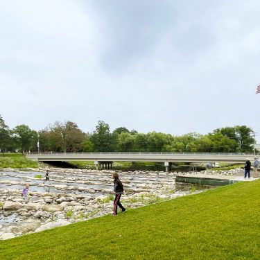Image-11 People walk along a grassy area beside a rocky stream, with a bridge, trees, and an American flag visible in the background under a cloudy sky.