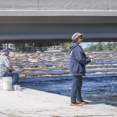 Image-12 Two people fish by a river, one sitting on a bucket and the other standing, both near a concrete bridge with rocks and flowing water visible in the background.