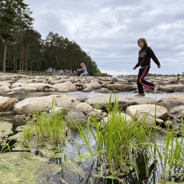 Image-9 Two girls are walking on large rocks across a shallow river, with green grass and algae in the foreground and tall trees in the background under a cloudy sky.